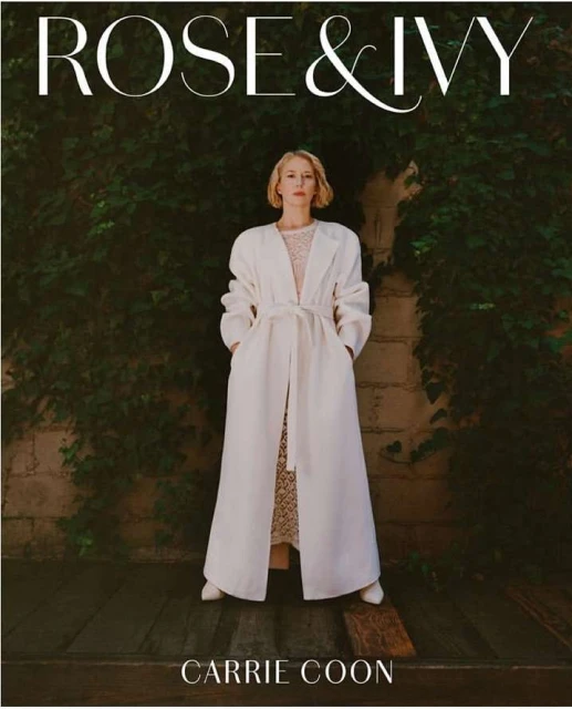 Carrie Coon poses in a long white coat, standing against a verdant ivy-covered wall, for Rose & Ivy, creating a striking portrait.