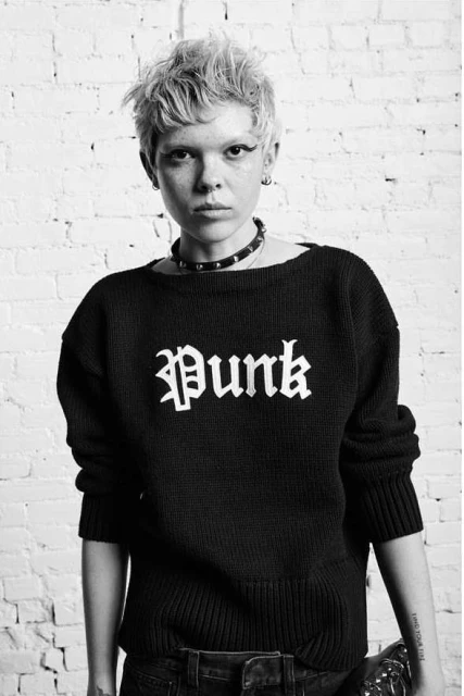 A young, alternative woman with short blonde hair in a black sweater with 'Punk' printed on the front, standing against a white brick wall.