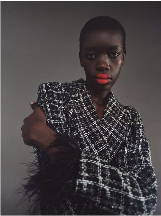Close-up of a Black woman with orange lipstick, wearing a plaid blazer with black feathered sleeves against a gray background.