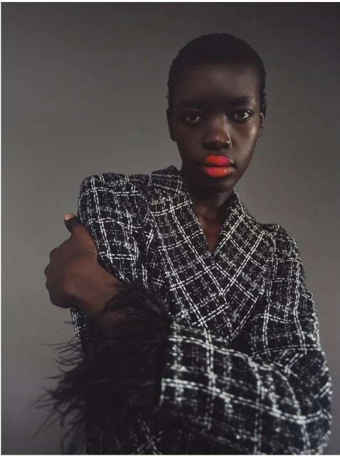 Close-up of a Black woman with orange lipstick, wearing a plaid blazer with black feathered sleeves against a gray background.