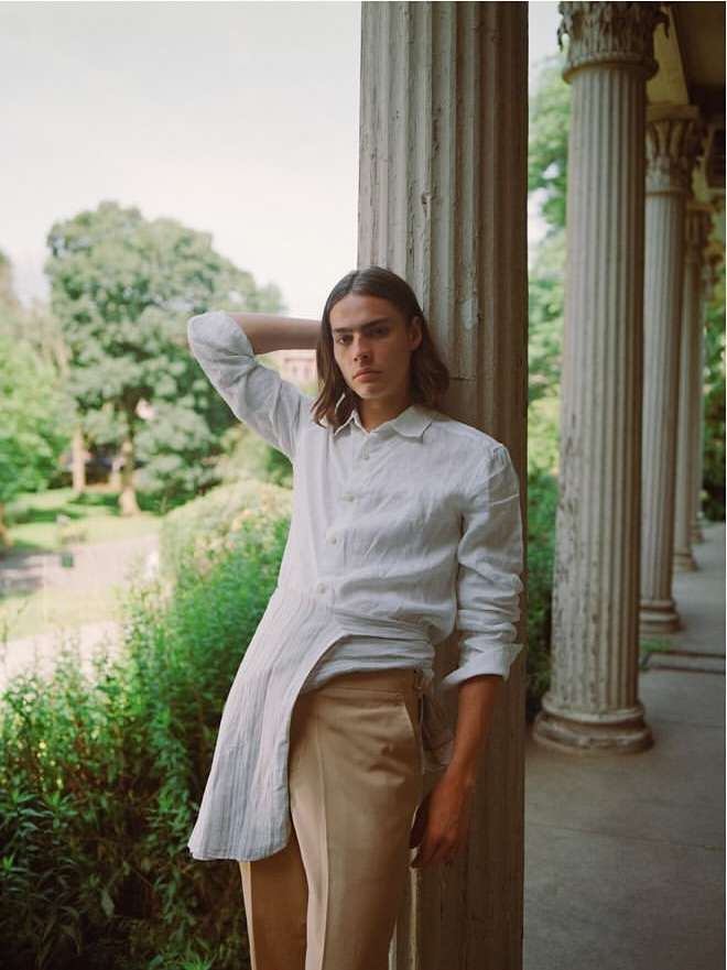 A young adult male posing outdoors in natural light. He is wearing a white button-down shirt and light brown pants, standing by stone columns.