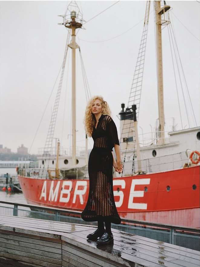 An adult woman with curly blonde hair poses on a wet wooden surface with a red boat behind her on an overcast day.