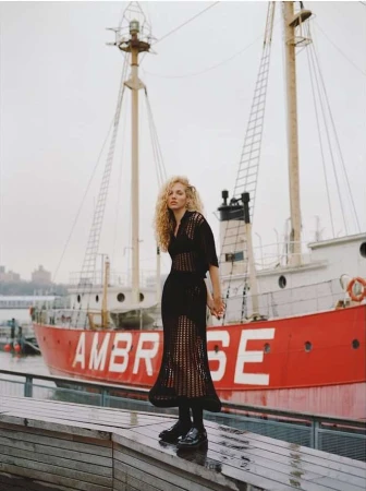 An adult woman with curly blonde hair poses on a wet wooden surface with a red boat behind her on an overcast day.