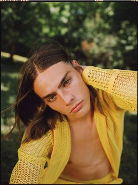 Portrait of a young adult male with long brown hair, fair skin, and a yellow knit cardigan, posing outdoors in a natural setting with green foliage.
