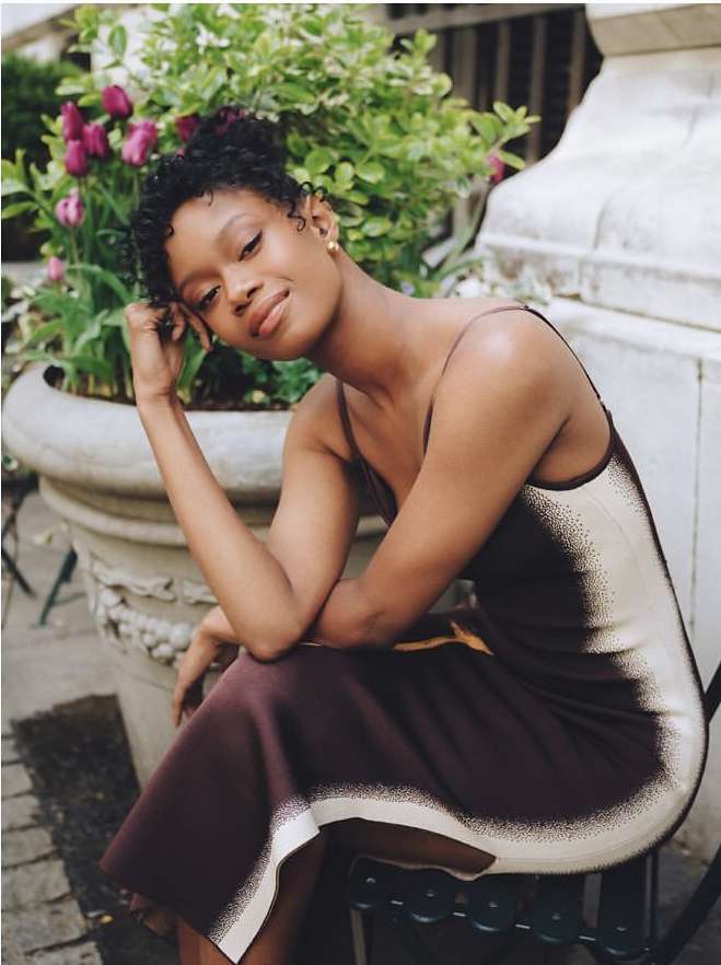 A young woman in an elegant brown dress poses outdoors near lush plants and stone planter, looking softly at the camera in a portrait.