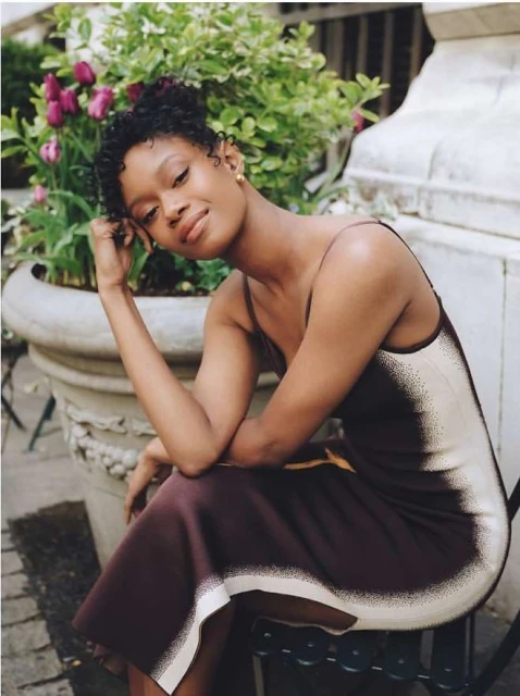 A young woman in an elegant brown dress poses outdoors near lush plants and stone planter, looking softly at the camera in a portrait.