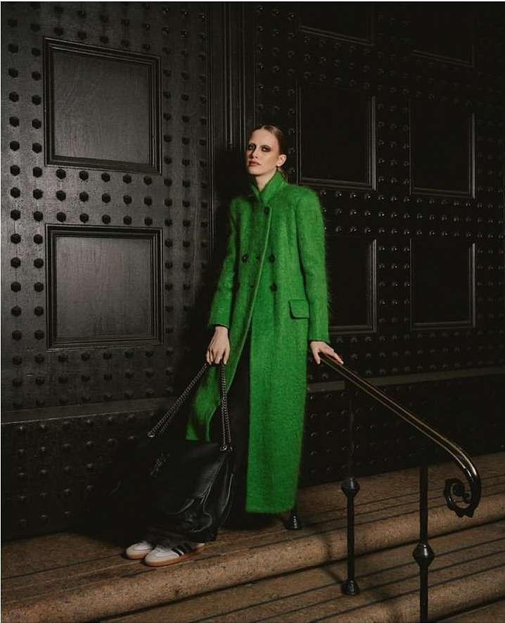 Model in a long green coat standing on stone steps holding a black leather handbag, photographed by Julia Sariy.