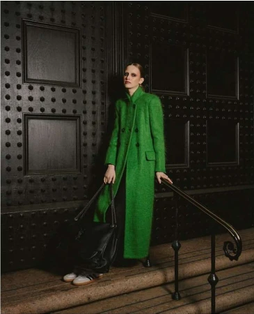 Model in a long green coat standing on stone steps holding a black leather handbag, photographed by Julia Sariy.