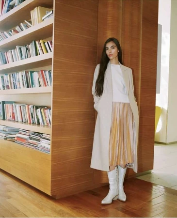 Woman in long cream coat, pleated skirt, and white boots standing beside a large wooden bookshelf wall.
