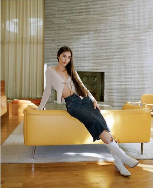 A woman sits on a yellow couch in a modern interior setting, wearing a white sweater, dark denim skirt, and white boots.