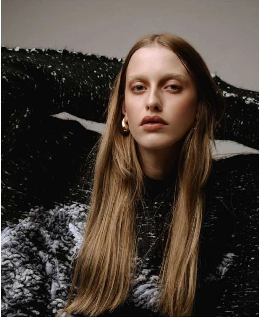 Portrait of a blonde-haired woman with gold hoop earrings and a textured top, posing against a black-and-white patterned backdrop.