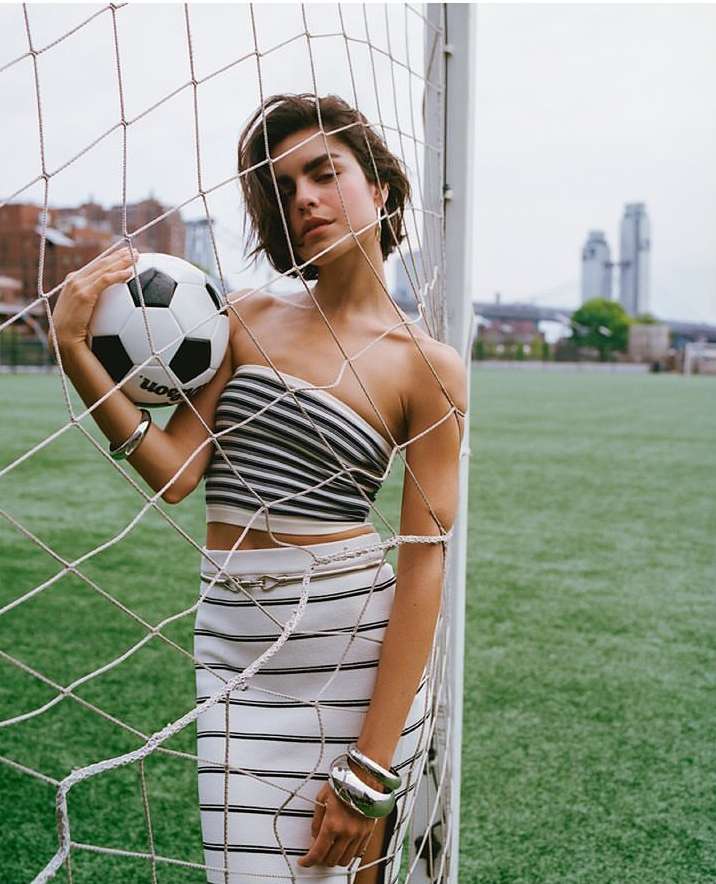 Woman posing with soccer ball in front of goal on green field, wearing striped top and skirt with silver jewellery, city skyline backdrop.