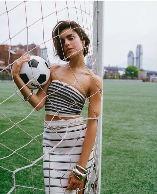 Woman posing with soccer ball in front of goal on green field, wearing striped top and skirt with silver jewellery, city skyline backdrop.