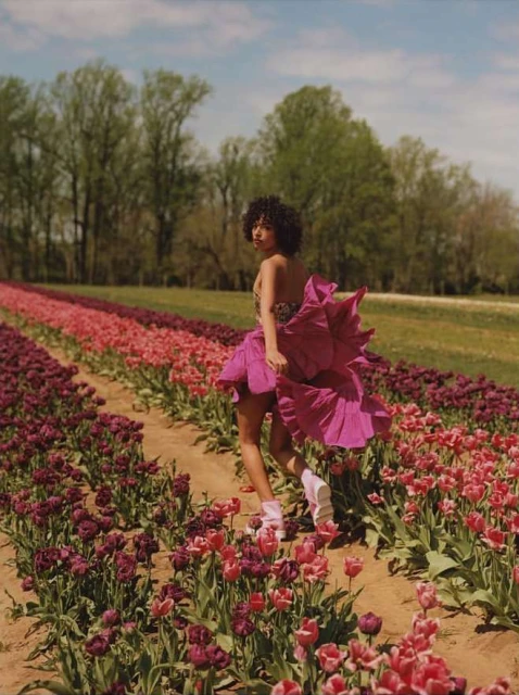A young woman in a purple dress walks through a field of colorful tulips on a sunny day with green trees in the background.