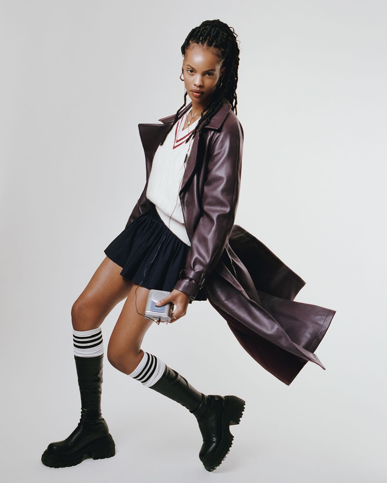 Stylish young woman in a burgundy trench coat, pleated skirt, and knee-high boots poses indoors while listening to music on a Walkman.