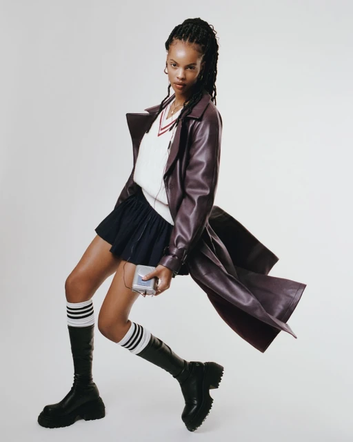 Stylish young woman in a burgundy trench coat, pleated skirt, and knee-high boots poses indoors while listening to music on a Walkman.