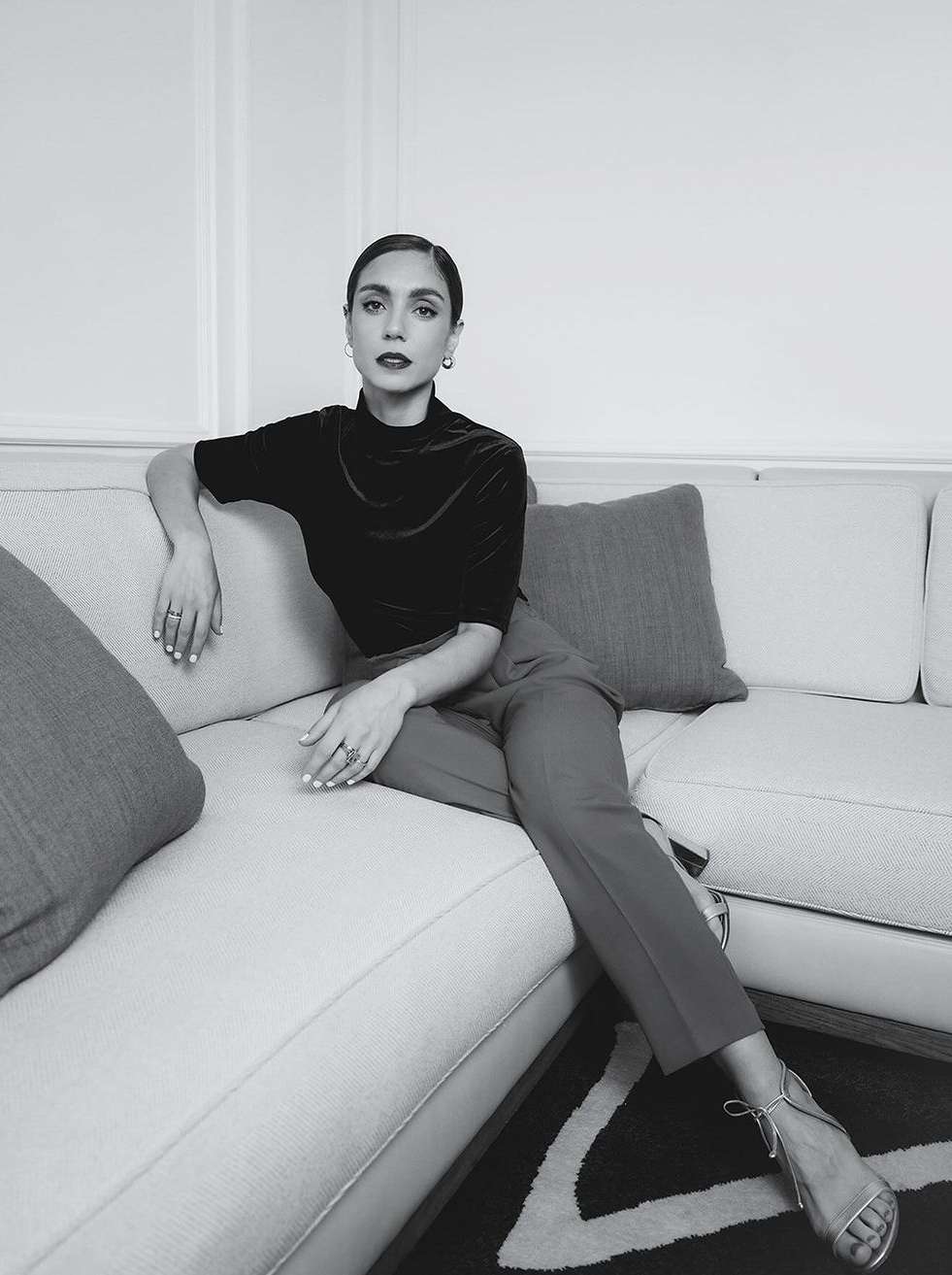 Black and white portrait of a woman with her legs crossed, sitting on a white couch with pillows and silver high heels.