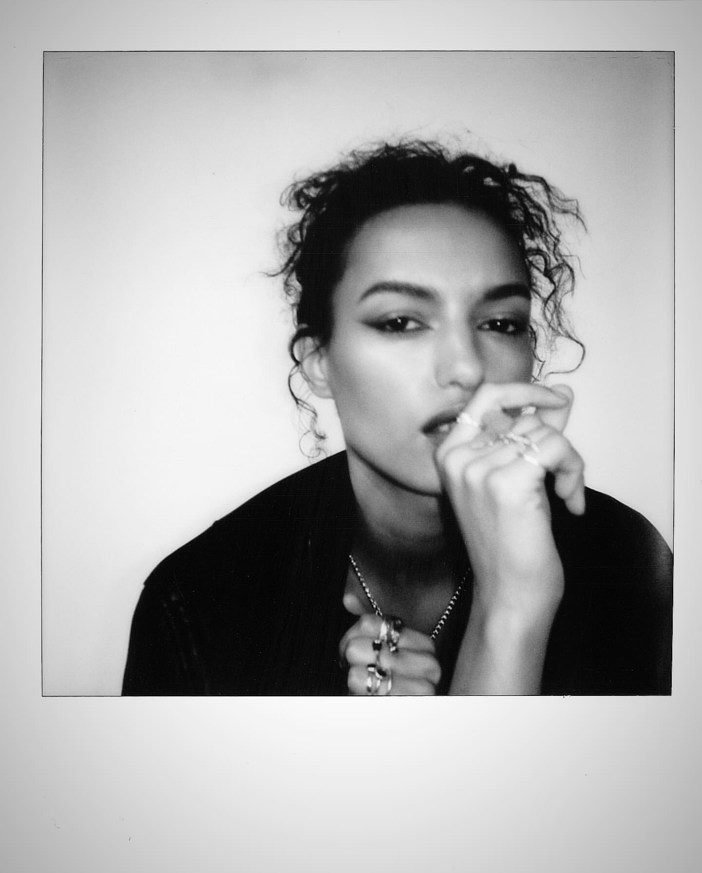 Portrait of a woman with curly hair in black and white, wearing rings and a necklace, with a hand near her face, in a close-up studio shot.