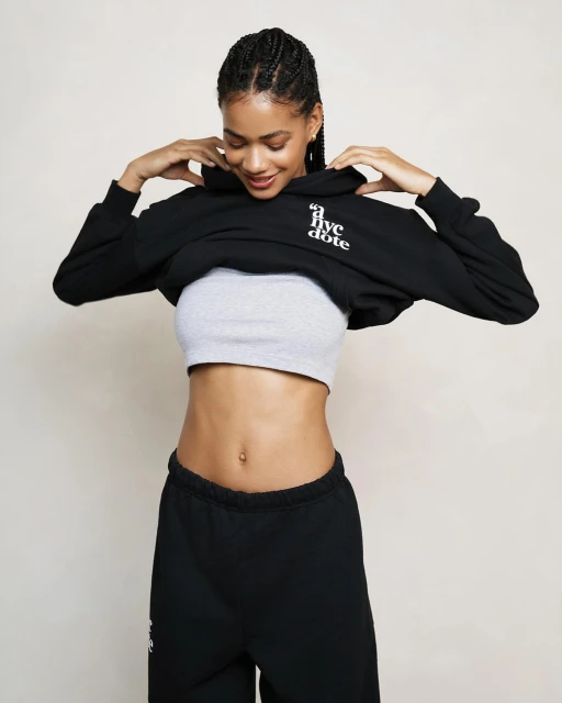 A smiling woman in a cropped black hoodie and grey top stands indoors in a bright setting, showcasing modern fashion.