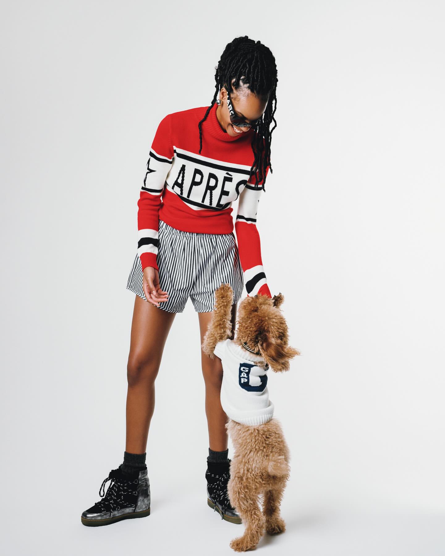 Woman in red sweater and striped shorts poses with a small dog in a white sweater on a seamless backdrop in a fashion shot.
