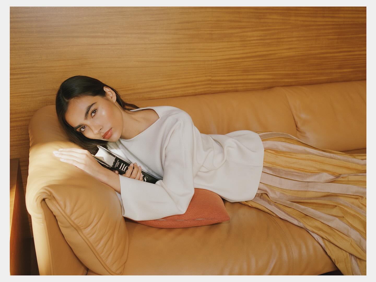 Brunette woman lying on a tan couch with a book, wearing a white long sleeve top and striped gold pants in an indoor setting.