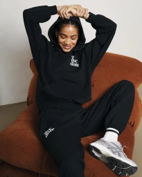 A woman in a black tracksuit and sneakers is seated in a large brown armchair, smiling with her hands clasped behind her head.
