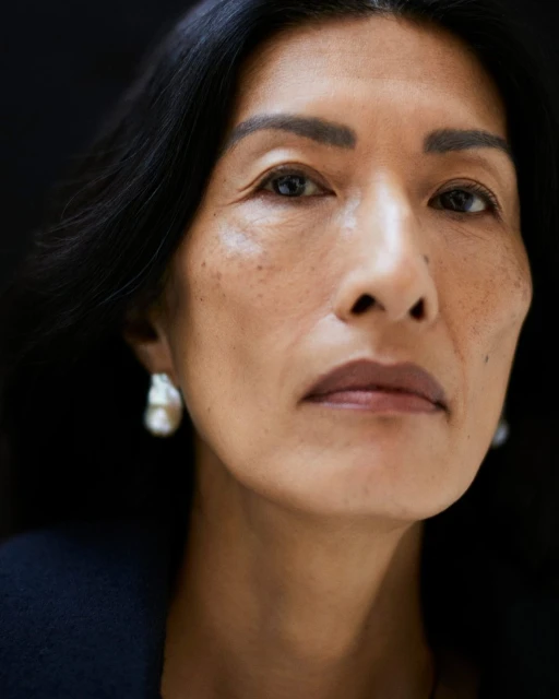 Close-up portrait of an adult female with dark hair, light skin, and pearl earrings against a dark background, showcasing beauty and elegance.