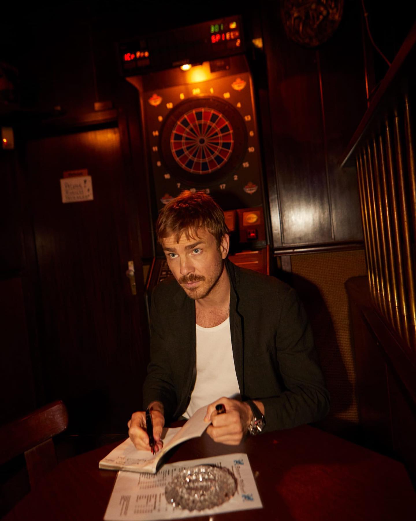 Indoor portrait of a man with a mustache sitting at a table writing in a notebook in a dimly lit bar with a dartboard in the background.