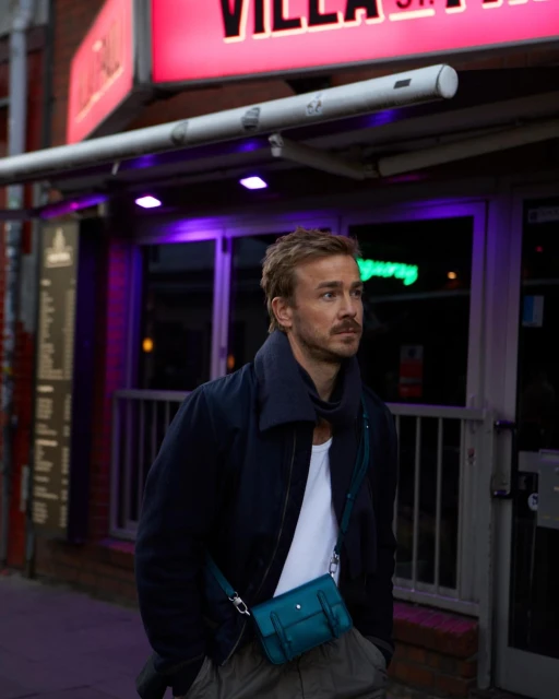 An adult man standing outdoors wearing a blue jacket, white shirt, and light pants, with a blue bag slung across his body.