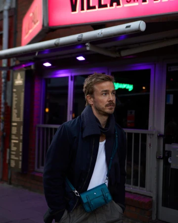 Man with mustache wearing a dark jacket and white t-shirt wearing a teal crossbody bag standing on a city street at night