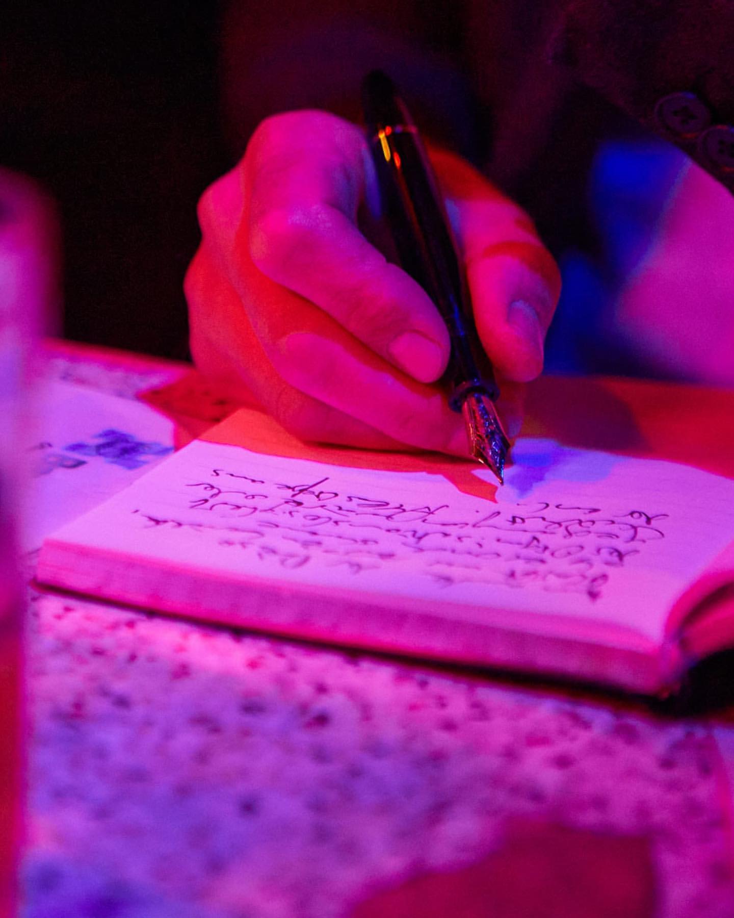 Close-up of a fountain pen writing on a notepad, with pink and purple lighting illuminating the hand, pen, and paper.