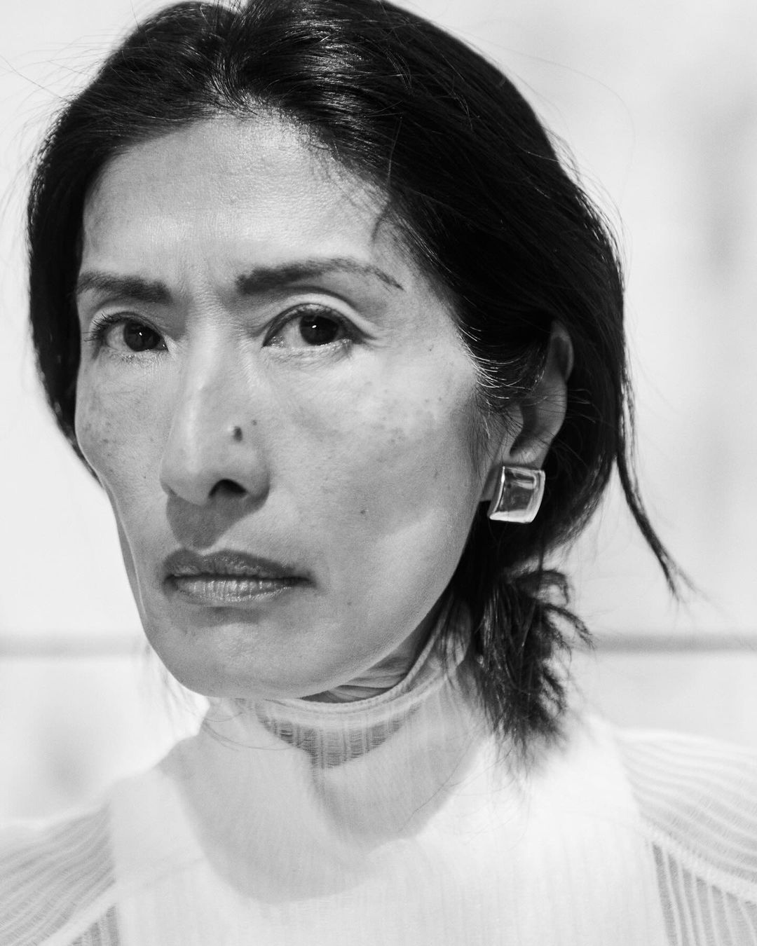 Black and white portrait of a woman with dark hair wearing earrings and a white top, looking directly at the camera with a neutral expression.