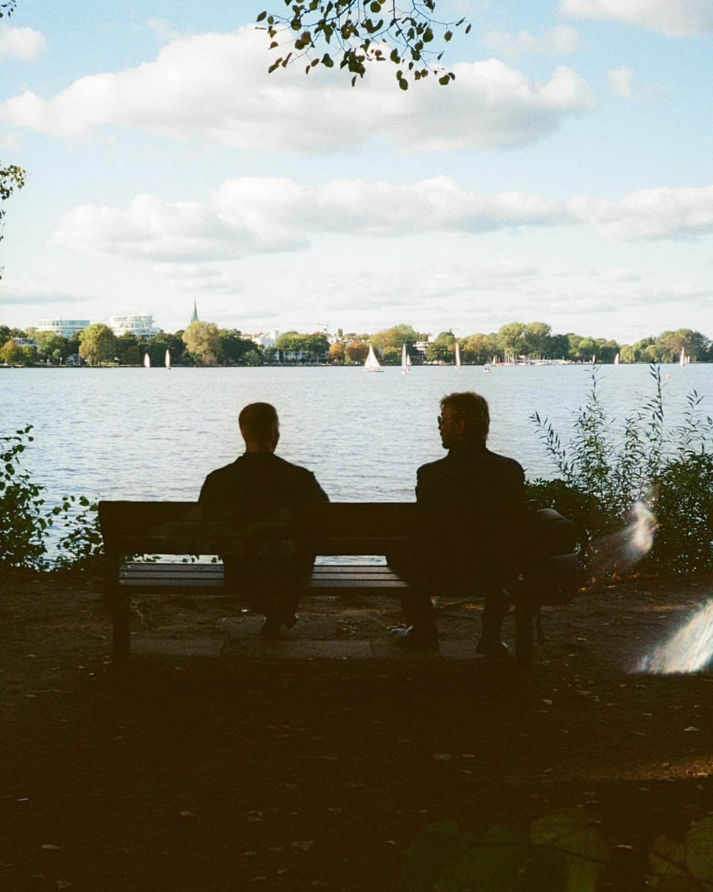 Two adult males sit on a bench looking at a lake with sailboats, under a cloudy sky. Silhouetted figures in a nature setting.