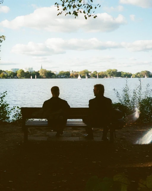 Two adult males sit on a bench looking at a lake with sailboats, under a cloudy sky. Silhouetted figures in a nature setting.