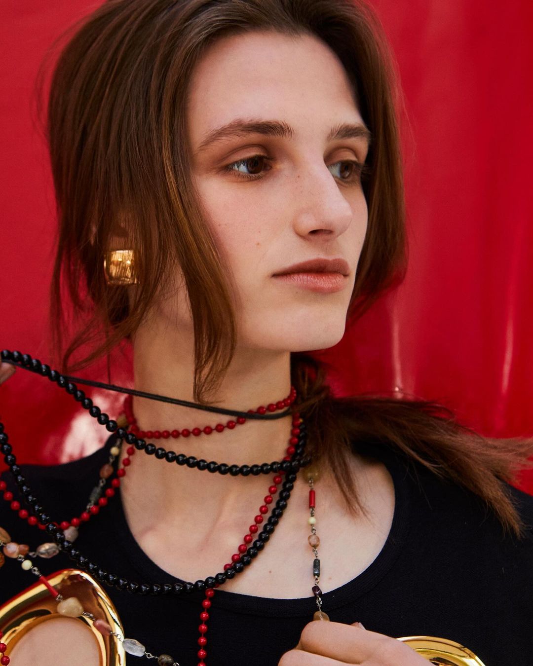 Fashion shot of a young woman with dark hair wearing gold earrings and multiple necklaces against a red background in soft, diffused lighting.
