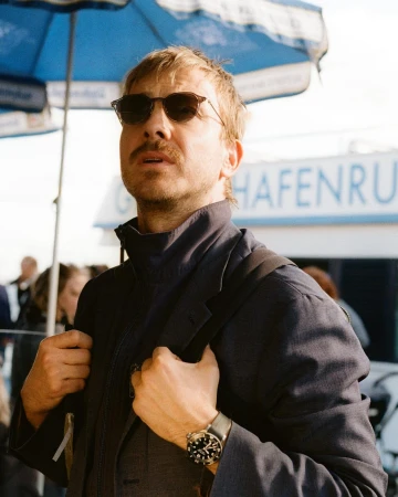 Man wearing sunglasses, mustache, and a dark blazer with a backpack strap, photographed outdoors by Julia Von Der Heide.
