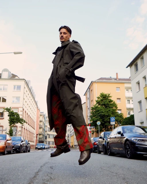A man in a trench coat mid-air on a city street wearing patterned pants and black boots, cars and buildings in the background.