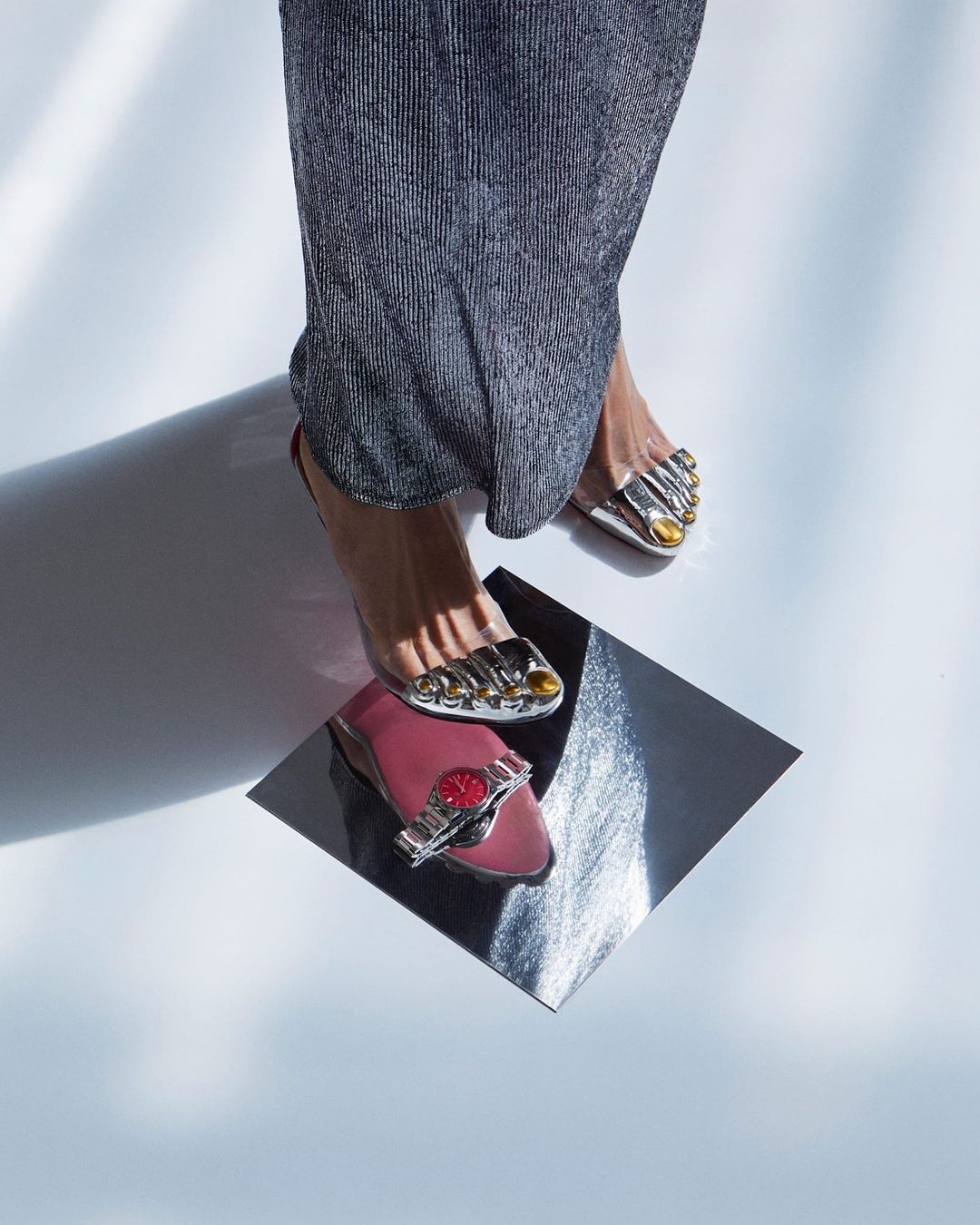 Elevated angle of feet in silver sandals on a reflective surface, pink flat with watch, metallic pleated fabric, and bright lighting.