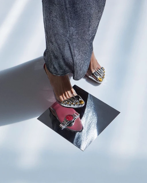 Elevated angle of feet in silver sandals on a reflective surface, pink flat with watch, metallic pleated fabric, and bright lighting.