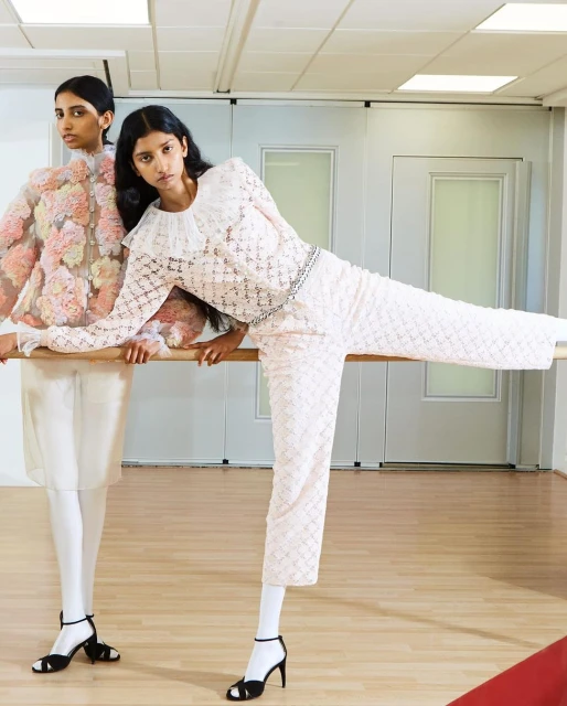 Two women pose in a ballet studio wearing couture fashion. One woman is in white pants and floral top, the other in floral coat.