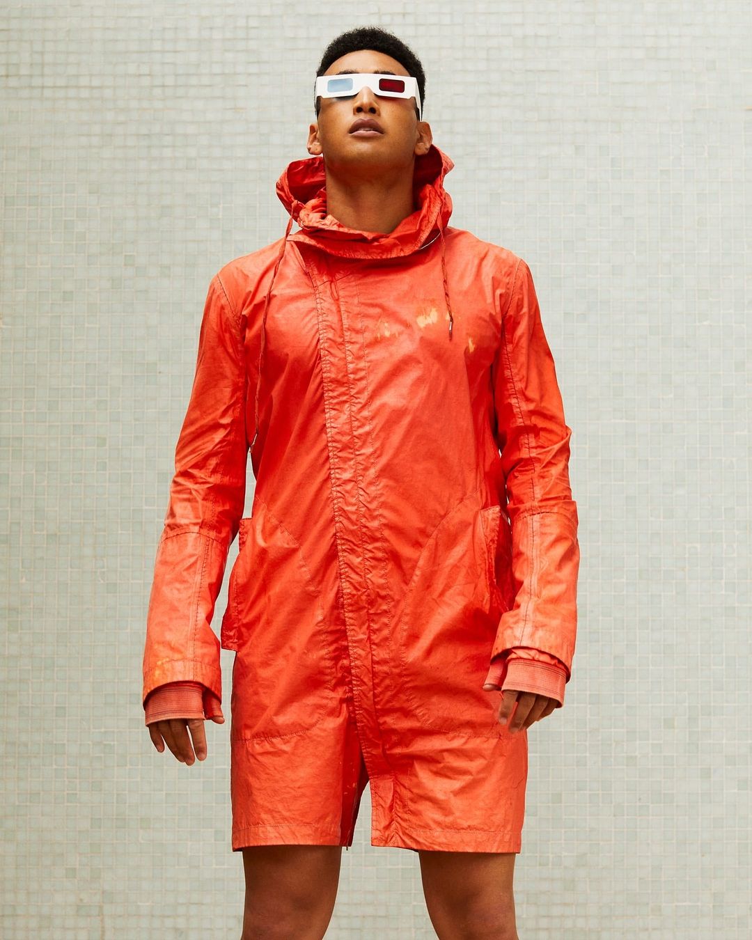 Man in an orange jacket and shorts wearing 3D glasses posing against a white tile wall in an eye-level medium shot with neutral lighting.