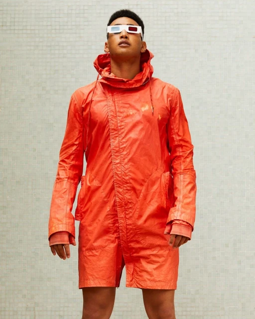 Man in an orange jacket and shorts wearing 3D glasses posing against a white tile wall in an eye-level medium shot with neutral lighting.