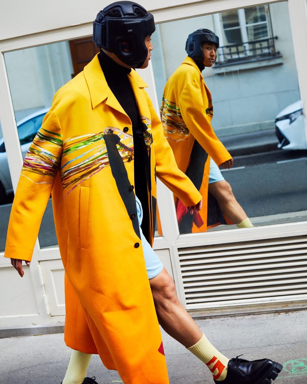 Man in yellow coat and black headgear walks past glass storefront, with reflection showing street scene and another view of the man.