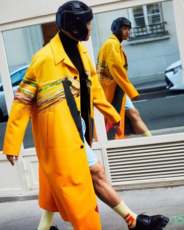 Man in yellow coat and black headgear walks past glass storefront, with reflection showing street scene and another view of the man.