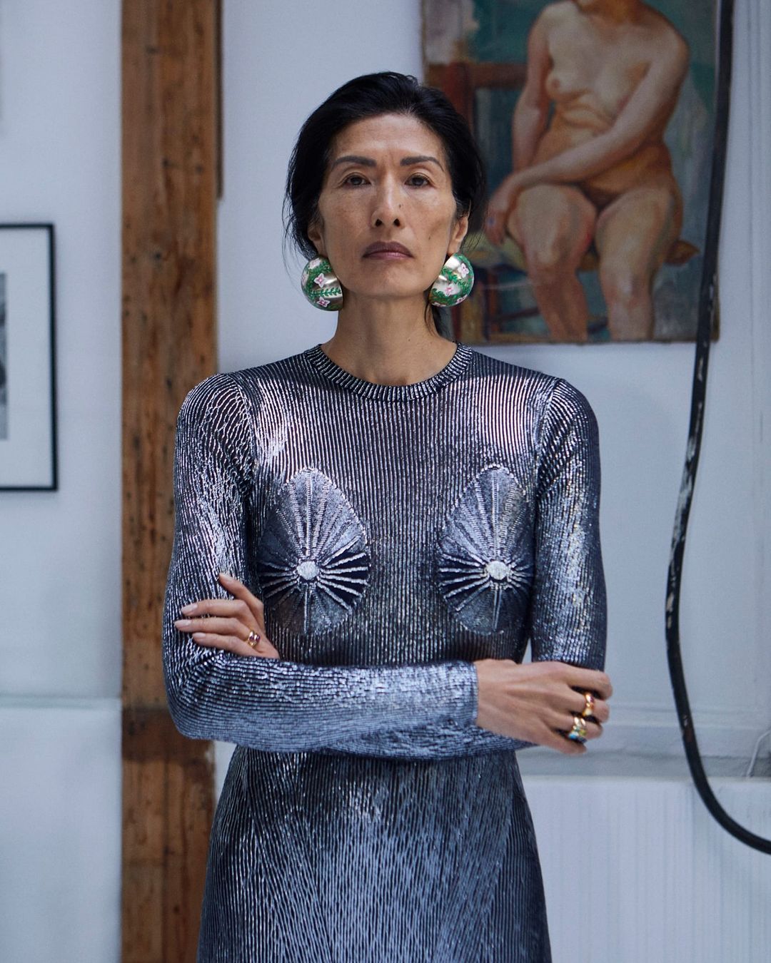 Elegant Asian woman in a silver dress with striking earrings stands in a bright indoor space with artwork visible in the background.