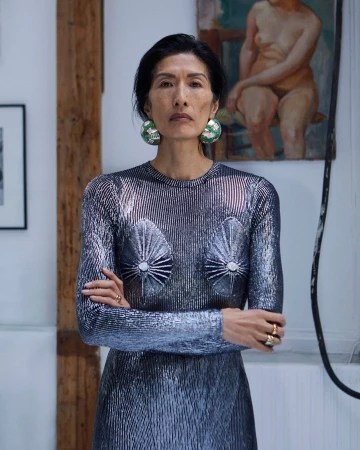 Elegant Asian woman in a silver dress with striking earrings stands in a bright indoor space with artwork visible in the background.