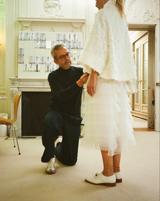 Fashion designer kneeling while fitting a white dress on a model indoors, surrounded by design boards and a formal interior setting.