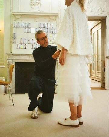 Fashion designer kneeling while fitting a white dress on a model indoors, surrounded by design boards and a formal interior setting.