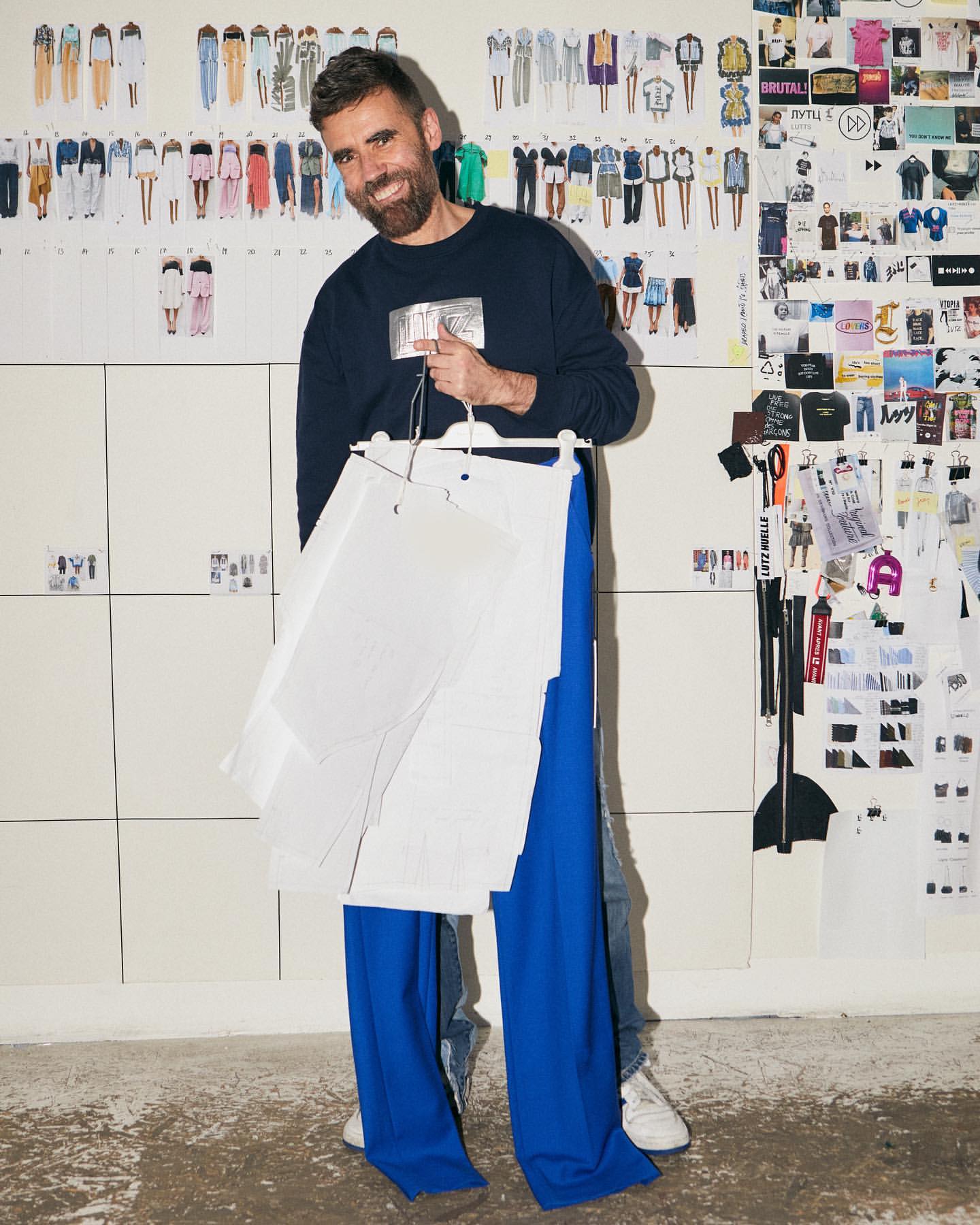 A man holding clothing patterns and blue pants with fashion design sketches on the wall, capturing a fashion design studio environment.