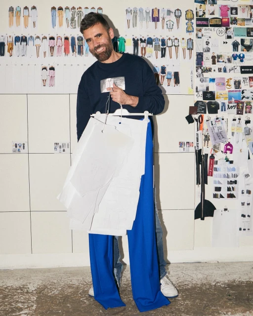 A man holding clothing patterns and blue pants with fashion design sketches on the wall, capturing a fashion design studio environment.